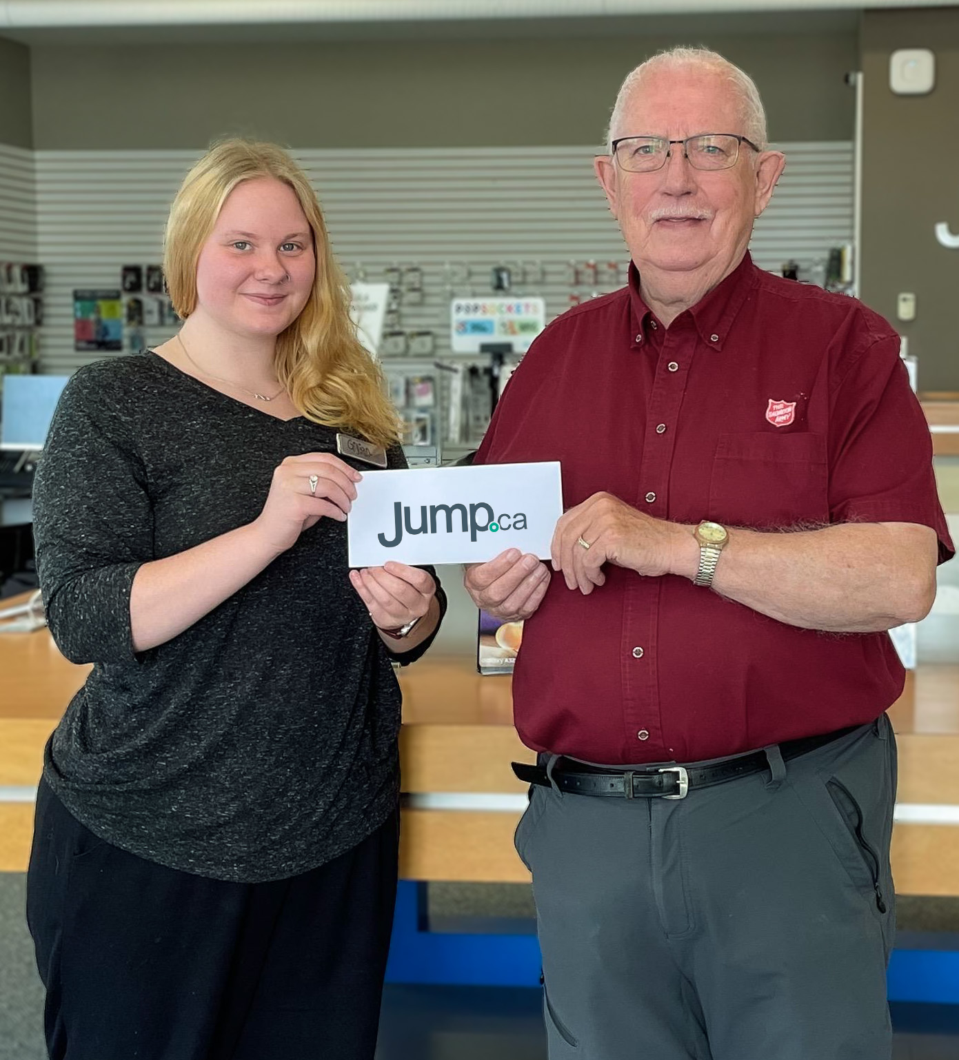 Two people, a young woman and an older man, smiling and holding a 'Jump.ca' sign inside a retail store with a background of phone accessories.