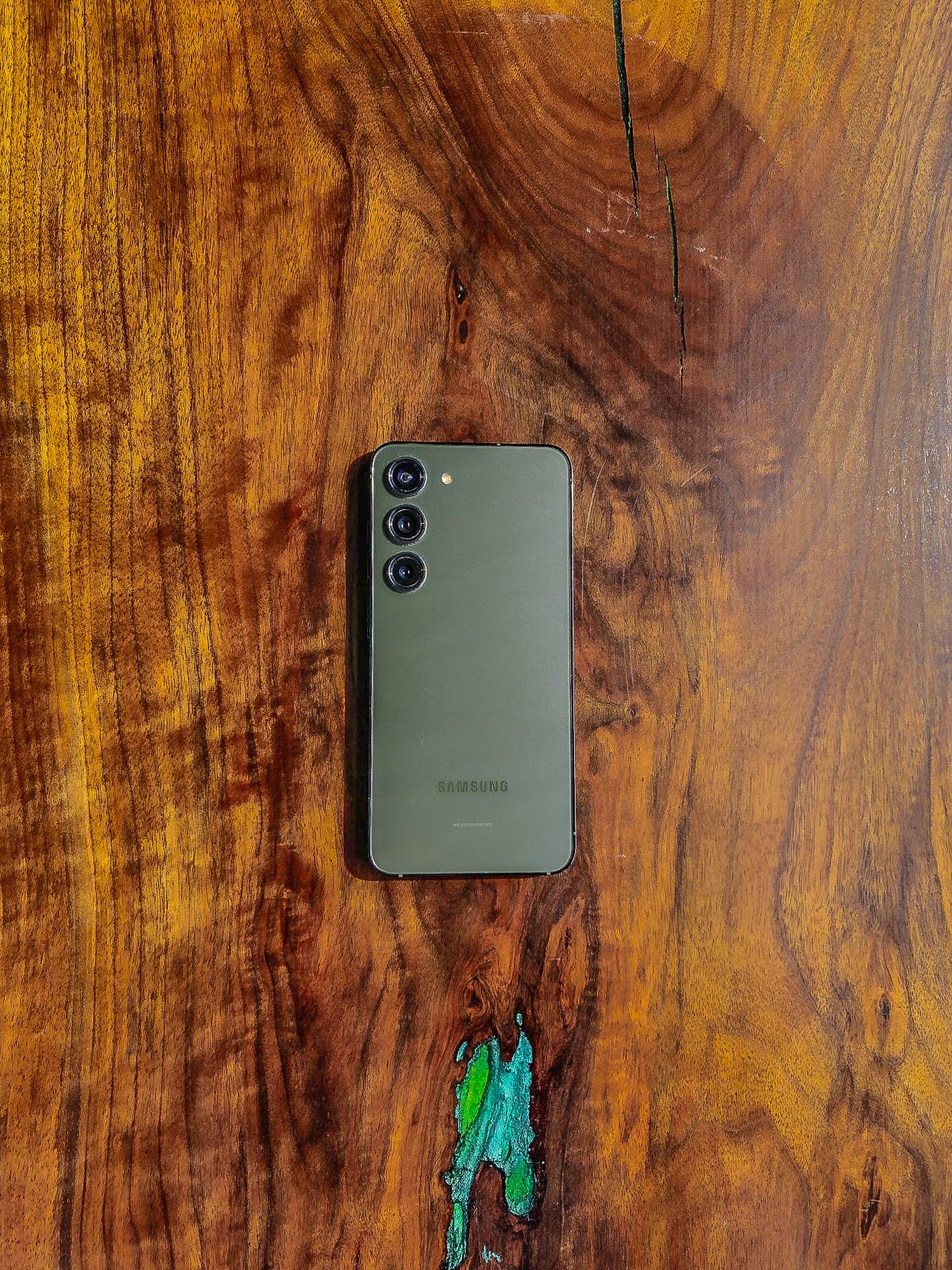Olive green Samsung smartphone with triple camera setup on a rich wooden background.